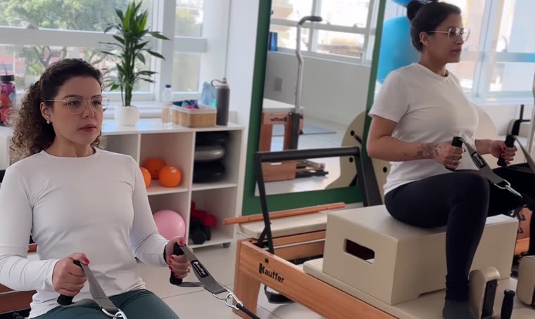 Duas alunas treinando ao mesmo tempo no estúdio Soliê Pilates, uma no reformer e outra na cadeira, cada uma com seu equipamento, numa aula de no máximo dois alunos por instrutora.