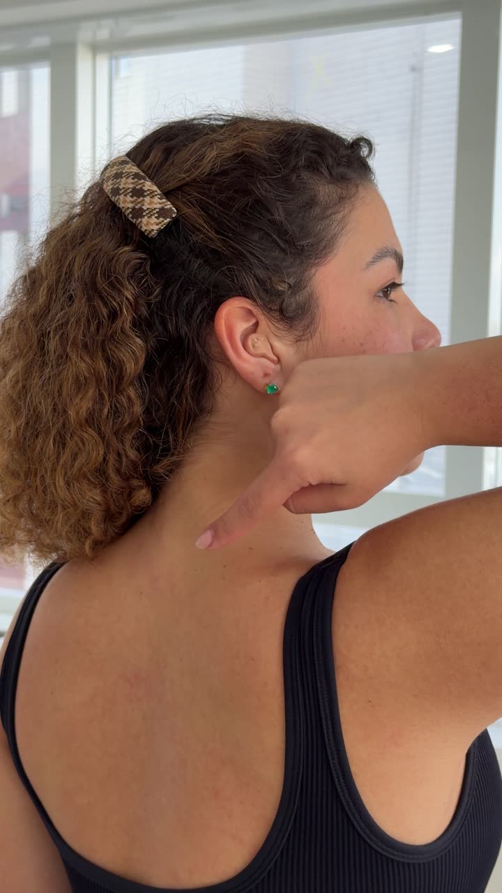 Vista de costas de uma mulher em regata preta apontando com o dedo indicador para a base do pescoço, na transição entre a coluna cervical e a coluna torácica, região onde costuma aparecer o volume conhecido como calombo na nuca.