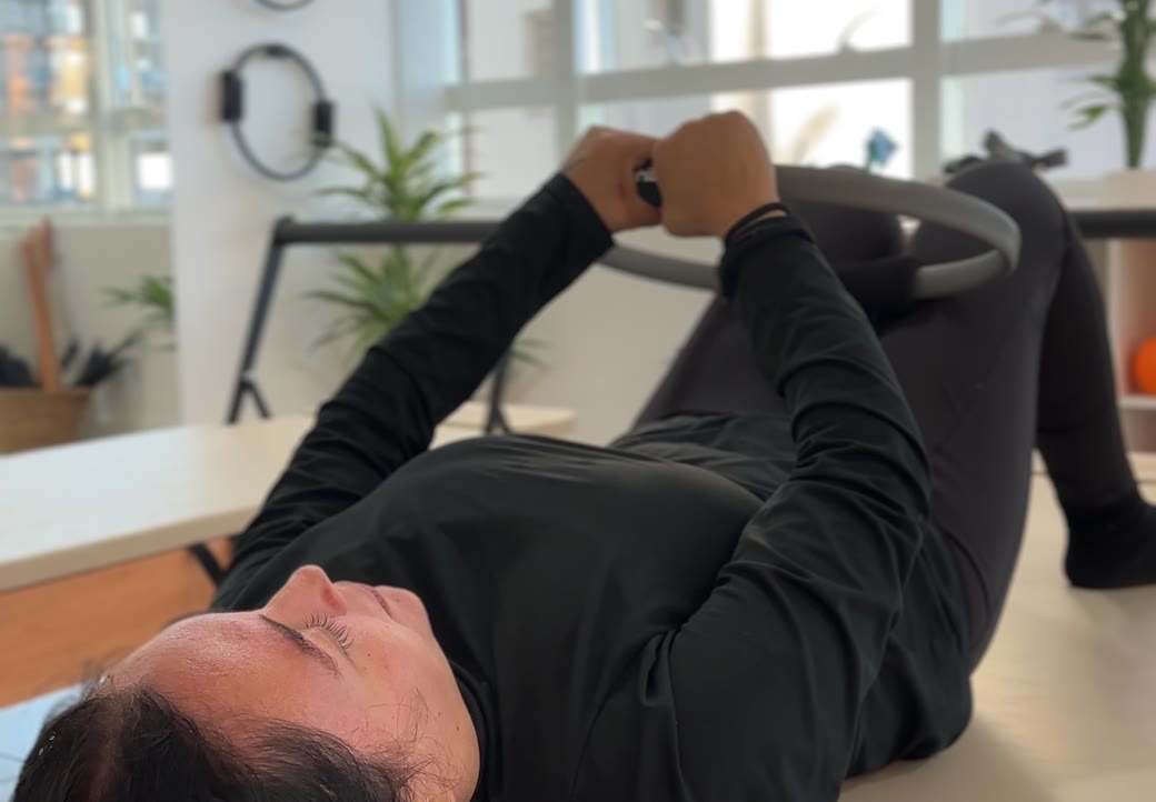 Ana, aluna da Soliê, deitada de costas sobre a maca do Pilates, joelhos flexionados, segurando um arco flexível (magic circle) com as duas mãos e os braços estendidos acima do peito. Trabalho de força de membros superiores e de core em amplitude controlada, sem impacto.