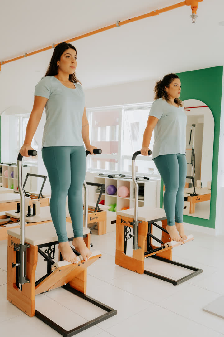 Duas instrutoras da Soliê em pé sobre os pedais de duas chairs de Pilates, segurando as barras laterais, trabalhando equilíbrio e força de pernas e tronco de forma simétrica