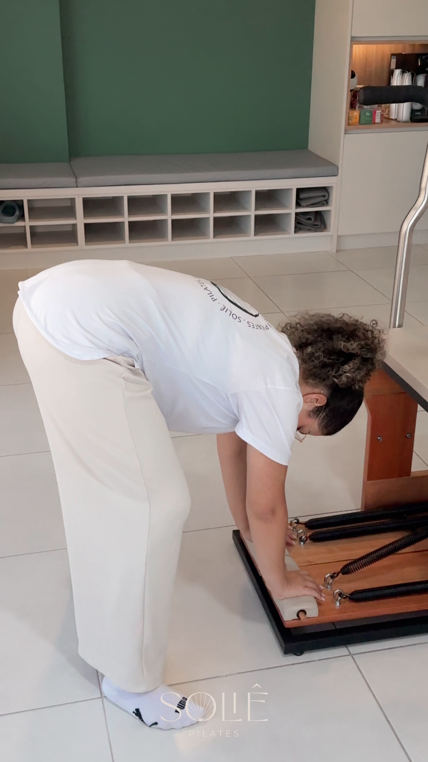 Gestante em pé inclinada à frente, com as mãos no pedal da cadeira de Pilates, fazendo o exercício de push down para alongar cadeia posterior