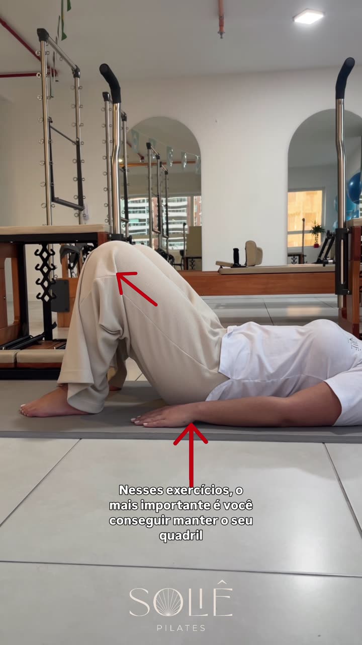 Instrutora da Soliê deitada de costas no tapete, joelhos dobrados e pés apoiados no chão, quadril elevado numa ponte de glúteo, com braços estendidos ao lado do corpo e as mãos pressionando o solo. Setas vermelhas indicam o quadril elevado e a mão apoiada no chão.