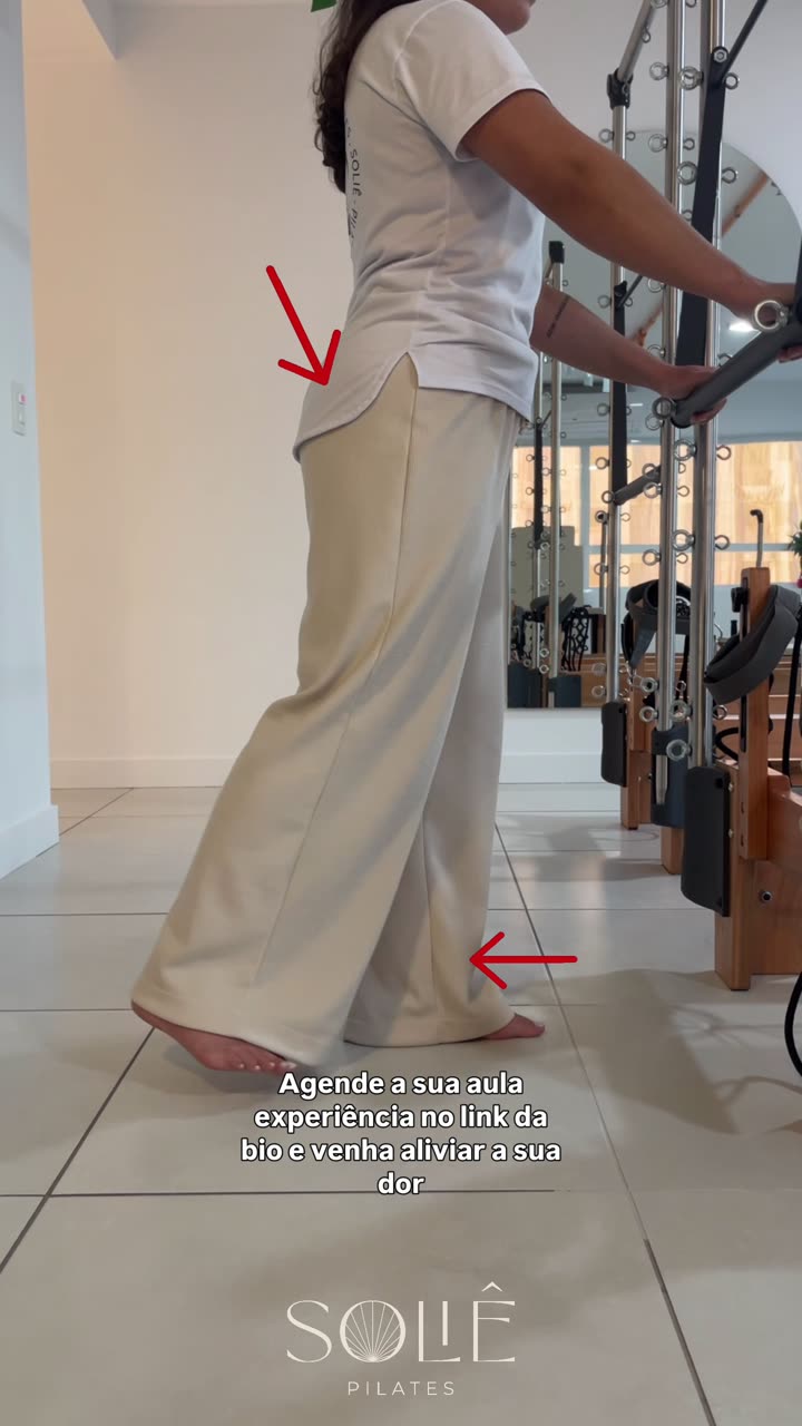 Instrutora da Soliê em pé de perfil, segurando um apoio vertical com as duas mãos, levando uma perna estendida para trás com apenas a ponta do pé tocando o chão, tronco ereto e pelve estável. Setas vermelhas indicam o glúteo contraído e a perna que vai para trás.