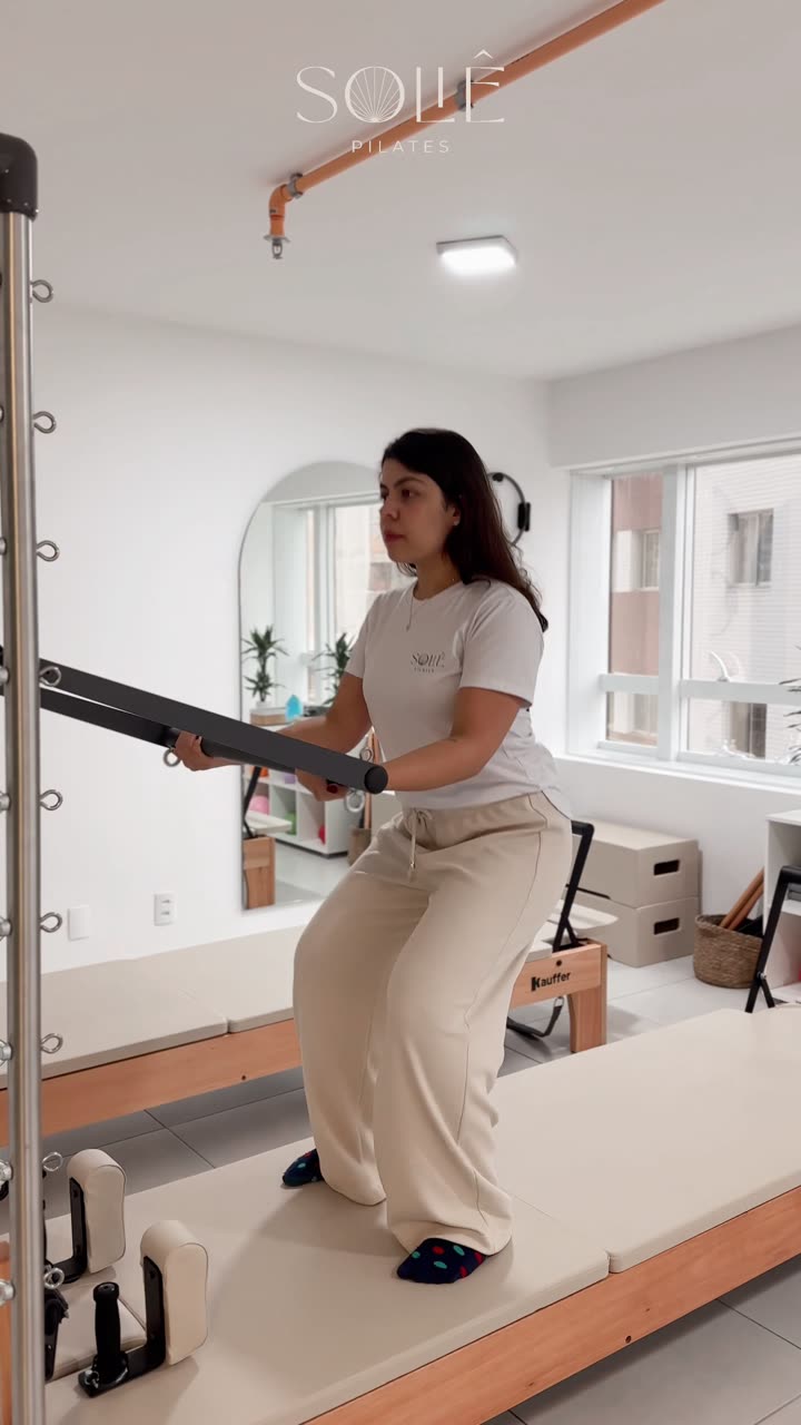 Aluna em pé sobre o reformer, segurando a barra do trapézio à frente, descendo em agachamento assistido para fortalecimento de quadríceps e glúteos