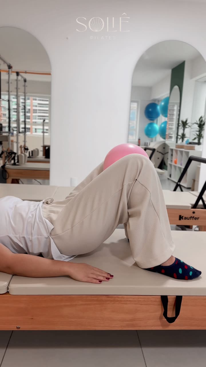 Aluna deitada de costas no banco do reformer, joelhos flexionados com uma bola pequena entre eles, fazendo elevação de quadril (ponte) para fortalecer glúteos e adutores