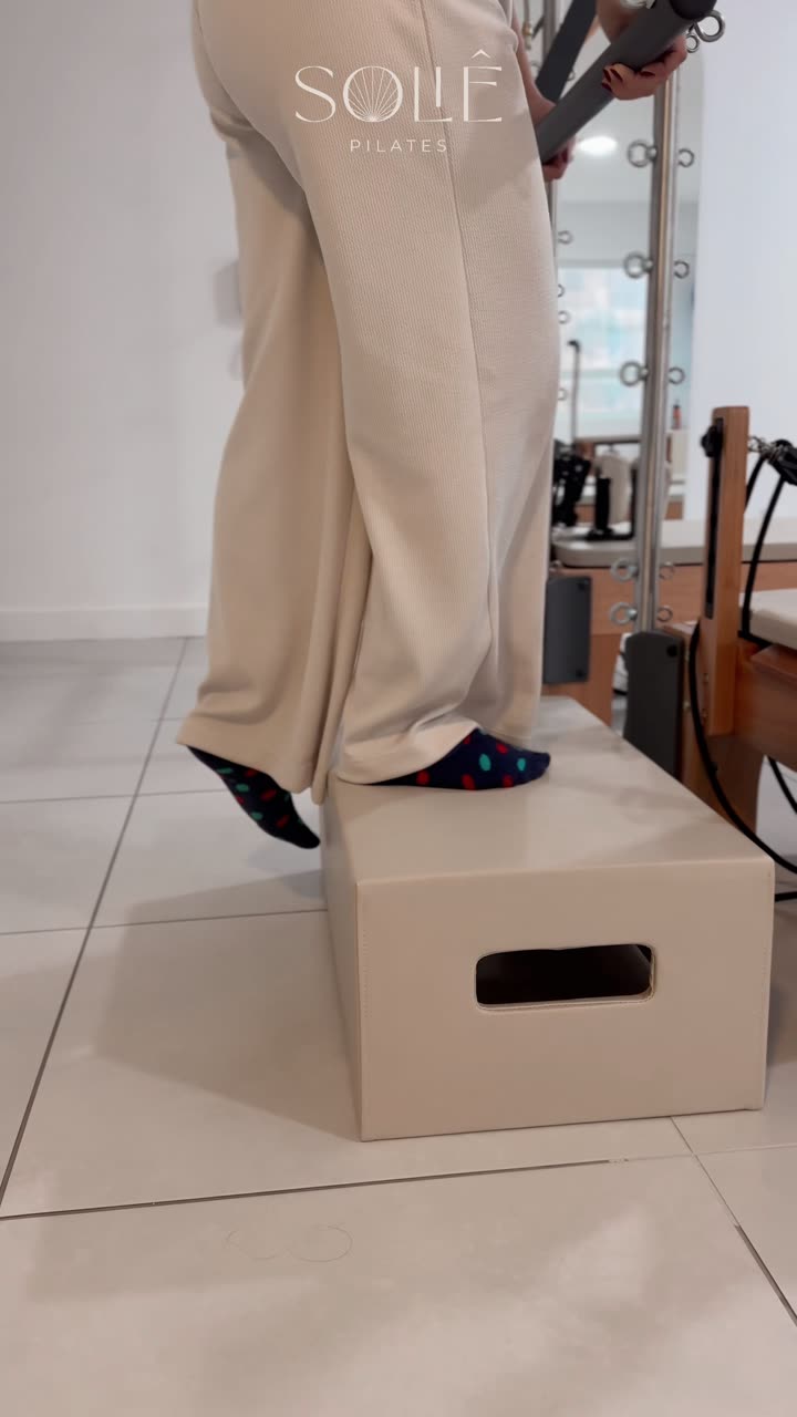 Aluna fazendo step up sobre um caixote do Pilates, com as mãos apoiadas na barra do trapézio, simulando o movimento de subir escada