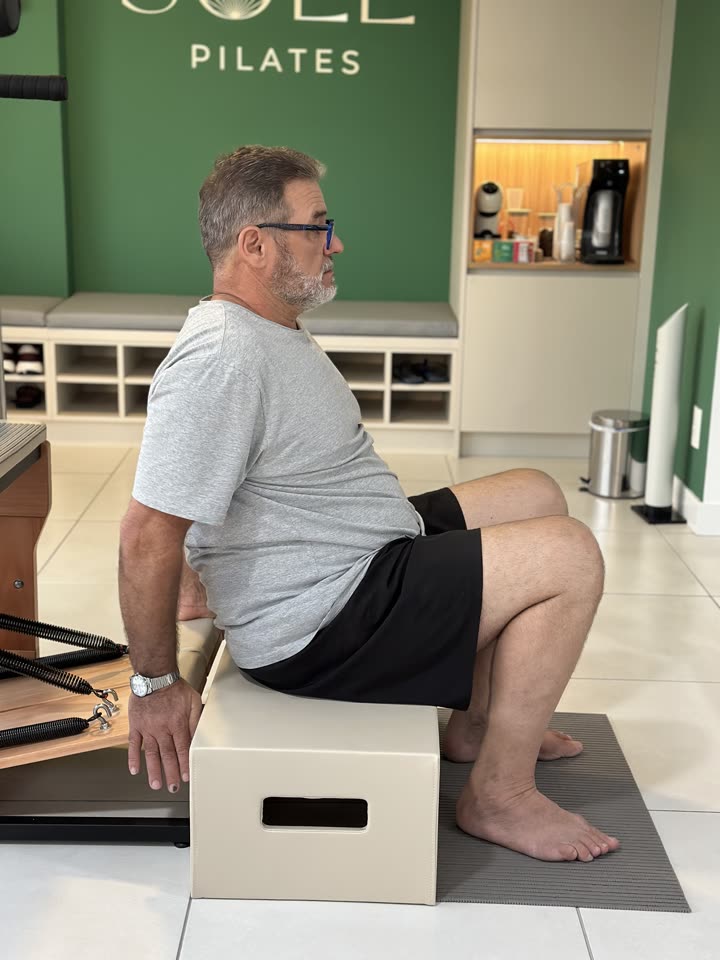 Aluno da Soliê Pilates sentado na borda de uma caixa de Pilates ao lado do Reformer, com os pés apoiados no chão e o tronco ereto, na posição de partida do movimento de sentar e levantar.