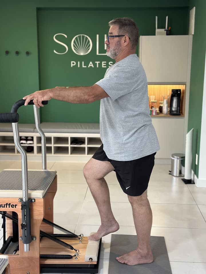 Aluno da Soliê Pilates em pé no estúdio, com as mãos apoiadas nas barras da Chair e um dos pés posicionado sobre o pedal do aparelho, iniciando o movimento de subida de degrau para fortalecer a perna de apoio.