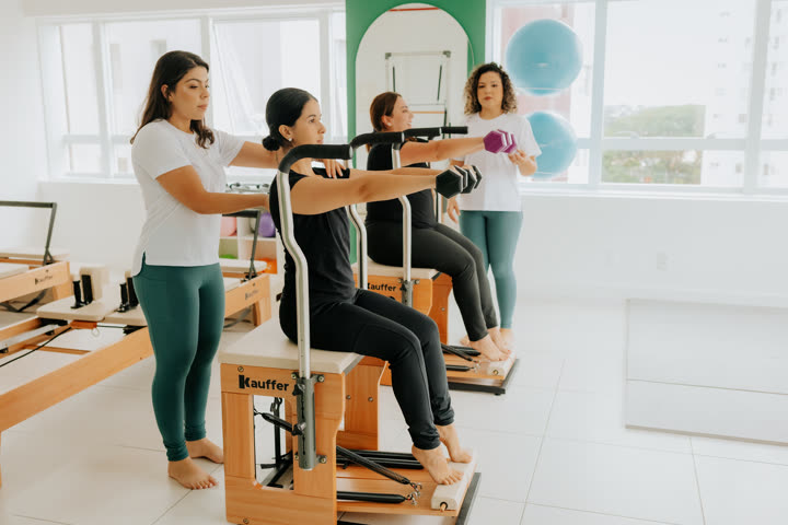 Duas alunas sentadas nas cadeiras de Pilates do estúdio Soliê fazendo elevação de braços com halteres, enquanto as instrutoras orientam o posicionamento dos ombros e das escápulas