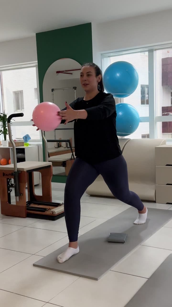 Aluna jovem da Soliê em afundo profundo sobre tatame, segurando uma bola overball entre as mãos com os braços projetados lateralmente. O exercício treina mobilidade do quadril, estabilidade do core e rotação controlada da coluna torácica.