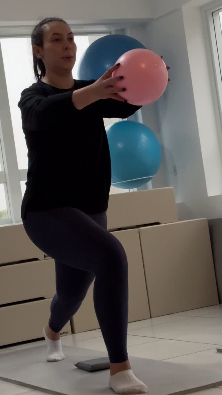 Aluna jovem da Soliê em apoio unipodal sobre tatame, joelho contralateral elevado, segurando uma bola overball à frente do tronco com os dois braços. Exercício de equilíbrio com controle motor fino, treinando estabilizadores profundos do quadril e do core.
