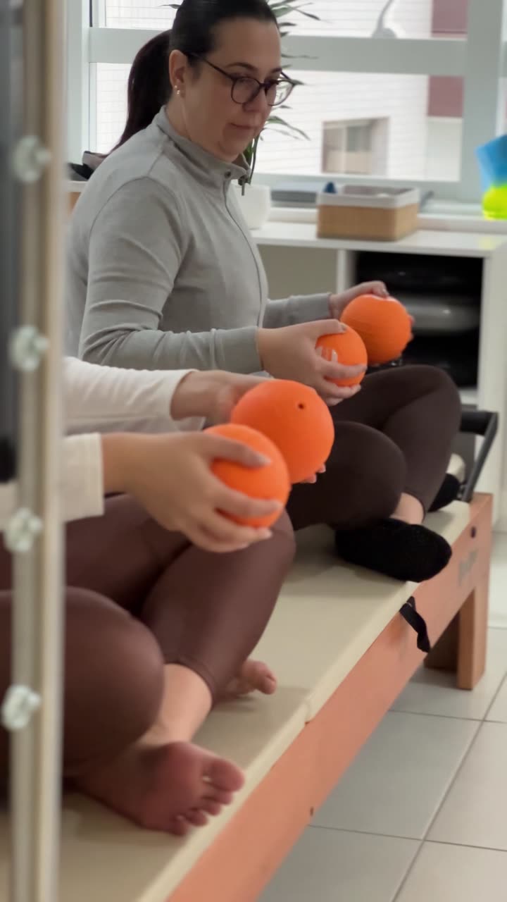 Aluna da Soliê sentada, segurando uma bola macia laranja entre as duas palmas das mãos à frente do peito, com os dedos espalhados sobre a bola e os punhos alinhados, comprimindo a bola num movimento de aperto que ativa a musculatura da mão e do antebraço.